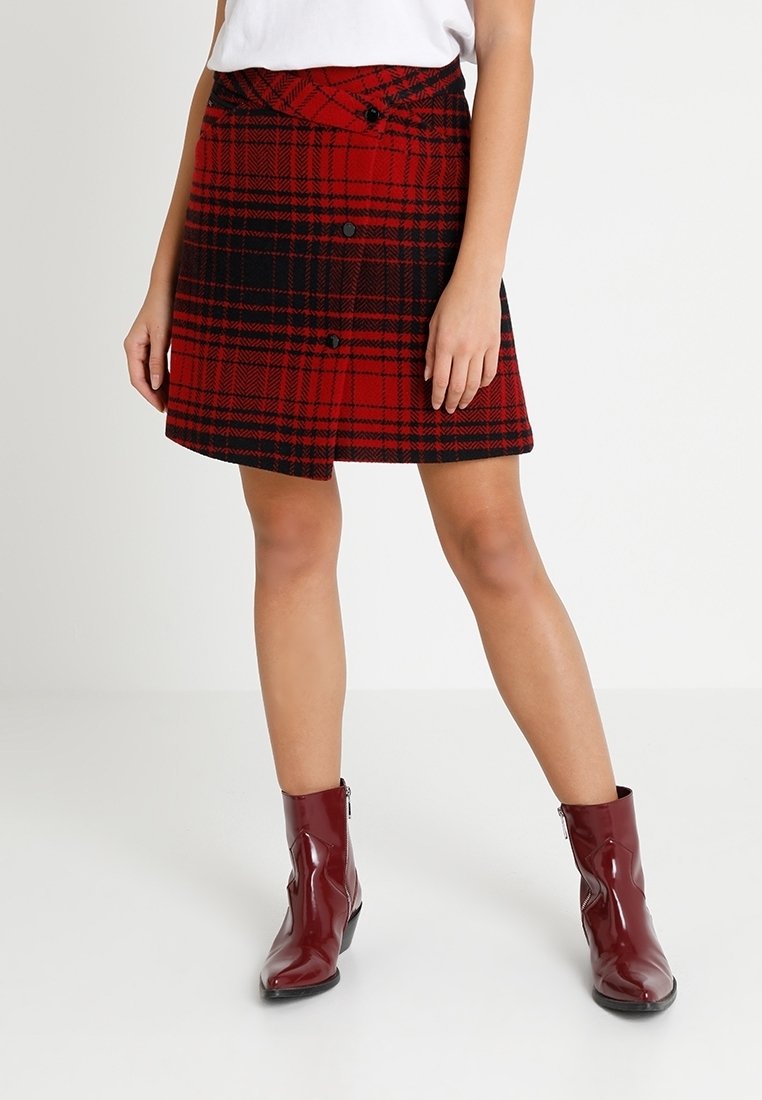 Femme portant une mini-jupe à carreaux rouge et noir avec des boutons et des bottines rouges brillantes, debout, les jambes écartées.
