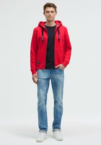 Sudadera roja con cremallera y cordones negros, con bolsillos frontales, llevada sobre una camiseta negra, combinada con jeans azul claro y zapatillas blancas.