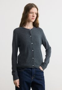 Γυναίκα με ανοιχτό καφέ μαλλιά, ντυμένη με γκρι σκούρο κουμπωτό cardigan και σκούρο μπλε τζιν, στέκεται με το ένα χέρι στην τσέπη.