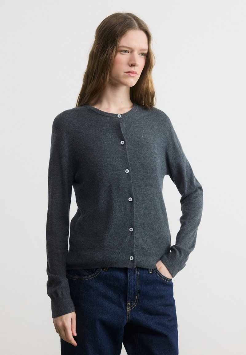 Γυναίκα με ανοιχτό καφέ μαλλιά, ντυμένη με γκρι σκούρο κουμπωτό cardigan και σκούρο μπλε τζιν, στέκεται με το ένα χέρι στην τσέπη.