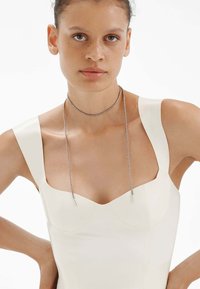 Collar de plata con un diseño delicado y texturizado; cuenta con acentos rectos. Llevado con una blusa blanca sin mangas y estructurada.