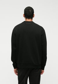 Man med kort mörkt hår klädd i en svart sweatshirt och byxor, stående med ryggen mot kameran mot en enfärgad bakgrund.