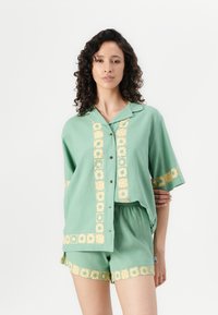 Lichtgroen katoenen twee-delige set bestaande uit een knoopsluiting met korte mouwen en bijpassende shorts, beide met gouden bloemenprintaccenten.
