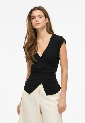 Kobieta z długimi brązowymi włosami, ubrana w czarny bezrękawnik w stylu kopertowym i szerokie spodnie o wysokim stanie w kolorze ecru na białym tle.