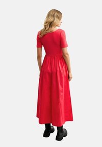 Red cotton maxi dress with short sleeves, fitted bodice, and gathered skirt. Worn with black chunky boots. Smooth texture, simple design.