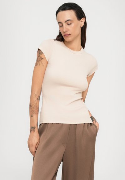 Donna con lunghi capelli scuri, tatuaggi sulle braccia, che indossa una maglietta beige aderente a maniche corte e pantaloni larghi marroni, in piedi con una mano in tasca.
