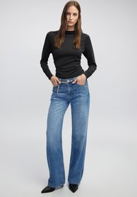Schwarzes Langarmoberteil kombiniert mit weit geschnittenen blauen Jeans, die mit Nietenakzenten und einer hellen Waschung versehen sind. Das Outfit wird durch schwarze spitze Schuhe ergänzt.