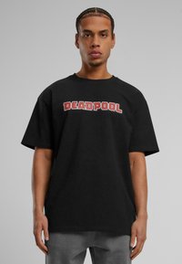 Black cotton t-shirt featuring bold "DEADPOOL" text in red letters, with a loose fit and short sleeves. Worn with gray jeans.