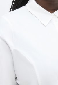 Κοντινό πλάνο ενός ατόμου που φοράει άσπρο πουκάμισο με γιακά, με μαλλιά πλεγμένα σε κοτσίδα να φαίνονται πάνω από τον ώμο.