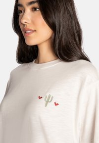 Camicetta bianca a maniche corte con ricamo di un cactus verde e un cuore rosso sul petto, realizzata in un tessuto morbido e leggero.