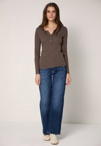 Braunes Henley-Oberteil mit langen Ärmeln, V-Ausschnitt und Knopfleiste, kombiniert mit blauen Jeans mit weitem Bein und weißen Turnschuhen.