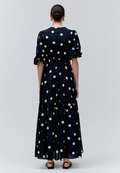 Kobieta z ciemnymi włosami upiętymi w kok, ubrana w granatową długą sukienkę w kropki z bufkami na rękawach oraz czarne sandały, stojąca na jasnym tle.