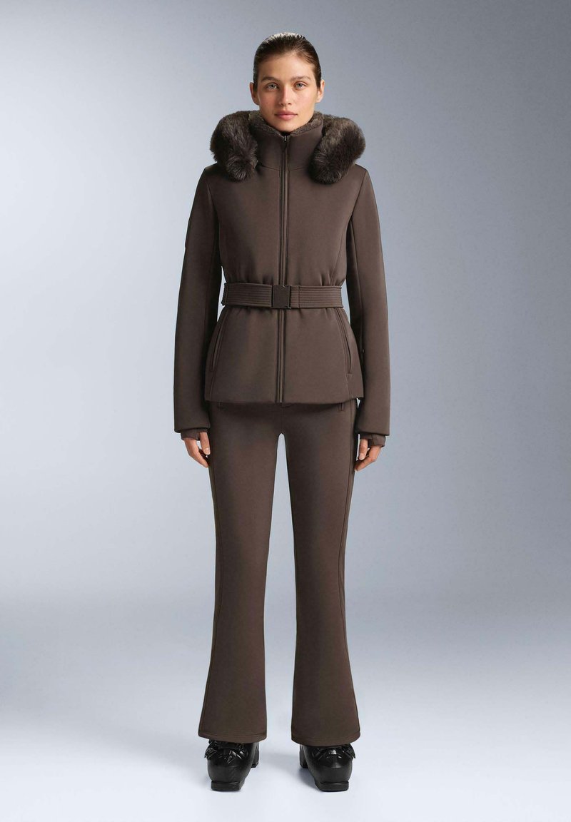 Hnědá, přiléhavá bunda s kožešinovým límcem, páskem v pase, dlouhými rukávy, v kombinaci s odpovídajícími přiléhavými kalhotami a lesklými černými botami. Hladká textura.