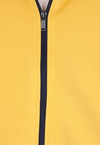 Giacca gialla con zip, caratterizzata da una cerniera blu navy, realizzata in un tessuto morbido e liscio con una superficie testurizzata. Vista ravvicinata del design.