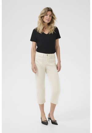 Schwarzes V-Ausschnitt-T-Shirt kombiniert mit cremefarbenen, knöchellangen Hosen. Die Hose hat frontale Taschen und ein figurnahes Design. Schwarze Absatzschuhe.