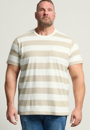 Mężczyzna z krótką brodą, ubrany w beżowo-biały, poziomo paskowany t-shirt z krótkim rękawem i niebieskie dżinsy, stojący na jednolitym tle.