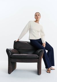 Vrouw in een wit shirt met lange mouwen en een donkere spijkerbroek zittend op de armleuning van een zwarte leren fauteuil tegen een effen witte achtergrond.