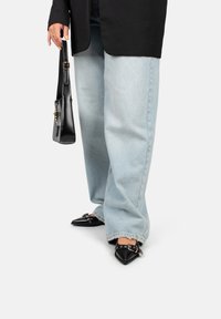 Pantalones de mezclilla de pierna ancha en azul claro, bailarinas negras de punta con detalles de hebilla y un bolso negro sostenido por una persona que lleva un blazer negro.