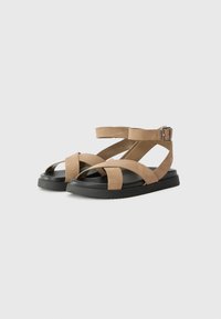 Sandalias de ante color topo con un diseño de tiras cruzadas, correa en el tobillo con hebilla y una suela de goma negra duradera. Forma plana y minimalista.