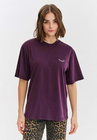 Fialové oversized tričko z bavlny s krátkými rukávy, s malou bílou výšivkou řas na hrudi. Nosené s kalhotami s leopardím potiskem.