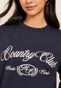 Sudadera azul marino con "Country Club Monte Carlo" bordado en crema, con un cuello redondo y textura suave. Pendientes de aro dorados visibles.