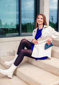 White oversized coat with large buttons, blue ribbed sweater dress, black tights, and white ankle boots. Holding a blue polka-dot clutch.