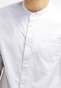 Camisa de manga corta, gris claro, con cuello tipo banda, botones al frente y un pequeño bolsillo en el pecho. Tejido texturizado con un brillo sutil.