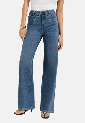 Woman wearing high-waist blue wide-leg jeans with tied belt, white sleeveless top, bracelet, and black heeled sandals.