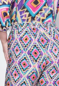 Byxor med elastisk midja som har ett livligt geometriskt mönster i lila, rosa, teal och guld, med en jämn textur och avslappnad passform.