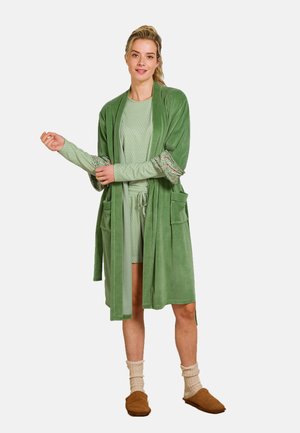 Groene velours badjas met zakken en gemusterde manchetten, gedragen over een lichtgroene outfit. Zachte textuur, relaxte pasvorm en gelaagd ontwerp.