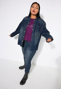 Chaqueta de mezclilla, lavado oscuro, cierre de botones, dos bolsillos frontales, combinada con una camiseta gráfica púrpura, jeans ajustados y botas negras de tobillo.