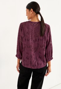 Lace blouse in donkerpaars met een gestructureerd bloemenpatroon, bootnek en driekwartmouwen. Gecombineerd met zwarte broeken.