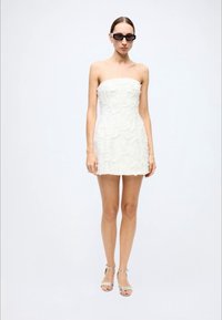 White strapless mini dress with a textured floral design. Model wears black sunglasses and silver ankle-strap heels. Smooth fabric finish.