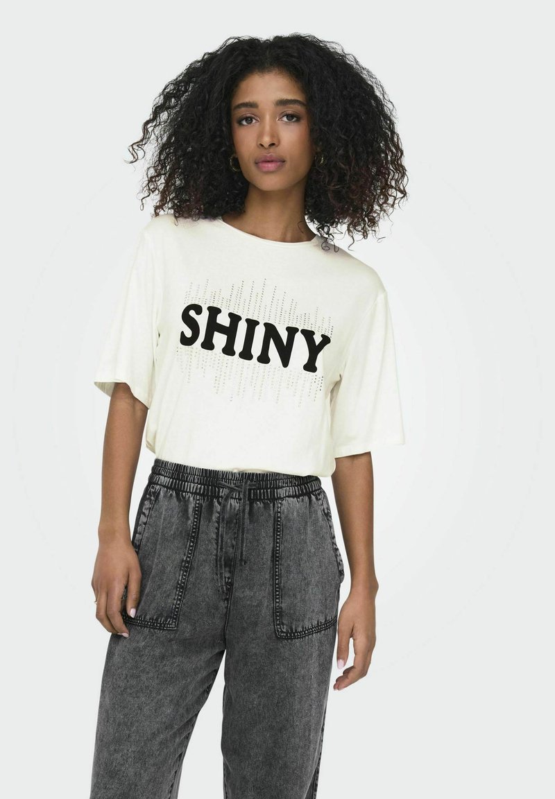 Cremefarvet t-shirt med korte ærmer og stort sort "SHINY" print samt sølvfarvede detaljer, parret med grå drawstring bukser med sidelommer.