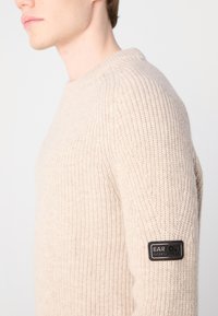 Beige bordás kötött pulóver hosszú ujjakkal és kerek nyakkivágással. A bal ujjon fekete Barbour logóval ellátott folt található.