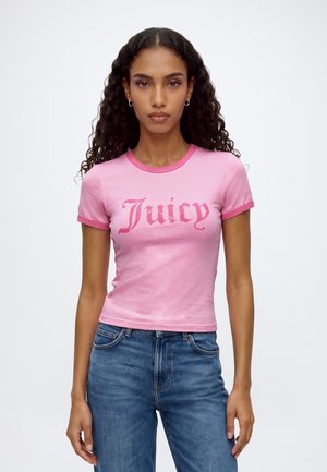 Femme aux longs cheveux bouclés portant un t-shirt court rose avec le texte "Juicy" et un jean bleu, debout devant un fond blanc uni.