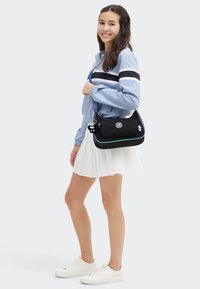 Giovane donna con giacca azzurra chiara, gonna bianca plissettata e sneakers bianche, che porta una borsa a tracolla nera con un accessorio a chiave.