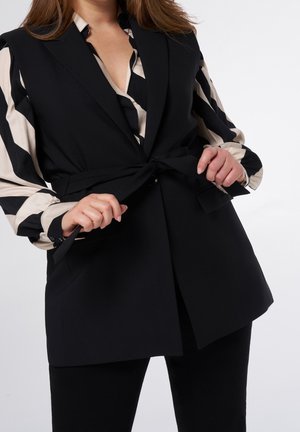 Kobieta wiążąca czarny pasek od bezrękawnika marynarki na czarno-beżowej, paskowanej, długorękawowej bluzce z czarnymi spodniami na białym tle.