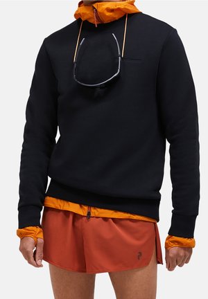 Mand iført sort langærmet sweatshirt over orange hættetrøje og orange løbeshorts, med solbriller hængende fra sweatshirtenes halsudskæring.
