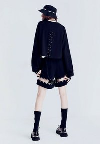 Suéter negro corto con espalda atada, combinado con pantalones cortos negros con herrajes decorativos y correas, complementado con un sombrero de cubo negro y calcetines.