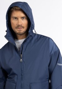 Giacca impermeabile blu navy con cappuccio, chiusura con zip e regolazioni elastiche. Presenta un logo sulla manica e tasche frontali. Texture liscia.