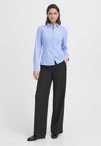 Világoskék csíkos, galléros gombos ing fekete bő szárú nadrággal és fekete mokaszin cipővel, fehér háttér előtt állva.