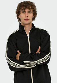 Schwarze Zip-Jacke mit hohem Kragen, mit cremefarbenen Streifen an den Ärmeln und einer glatten Textur. Erhältlich in lockerer Passform.