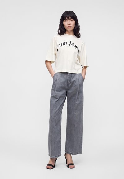 Γυναίκα που φοράει άσπρο oversized t-shirt Palm Angels, γκρι τζιν με φαρδιά μπατζάκια και μαύρα σανδάλια με τακούνια, στέκεται με τα χέρια στις τσέπες.