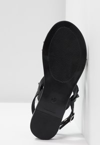 Sandalía negra con suela plana y textura antideslizante, cierre de hebilla y tiras delgadas, que exhibe un diseño minimalista.