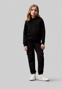 Felpa nera con cappuccio, abbinata a pantaloni cargo neri e sneakers bianche. La felpa presenta un piccolo logo sul petto.