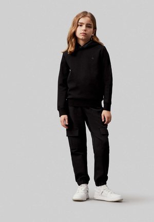 Felpa nera con cappuccio, abbinata a pantaloni cargo neri e sneakers bianche. La felpa presenta un piccolo logo sul petto.