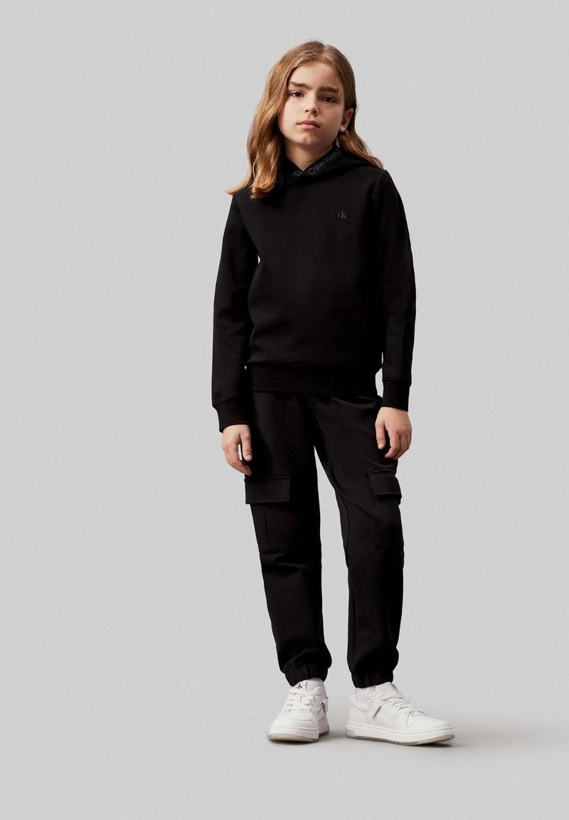 Felpa nera con cappuccio, abbinata a pantaloni cargo neri e sneakers bianche. La felpa presenta un piccolo logo sul petto.