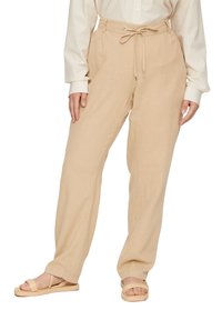 Beige linnen broek met een relaxte pasvorm, een trekkoord in de taille en zijzakken. Draagt met lichtgekleurde platte sandalen.