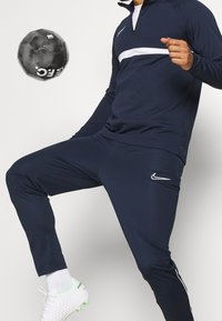 Granatowa sportowa bluza dresowa z białym paskiem na wysokości klatki piersiowej oraz spodnie do kompletu; posiada logo Nike i swobodny krój. Białe korki z neonowymi akcentami.